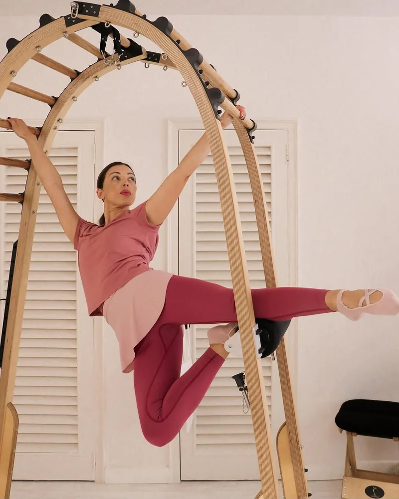 Laura Lojo, fondatrice di Art Pilates, in una posa dinamica appesa a un grande arco di legno in uno studio luminoso
